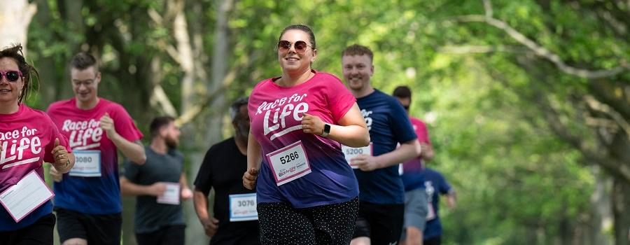 A group of runners wearing Race for Life T-shirts and sunglasses as they take part in a 5K race in a park on a sunny day.