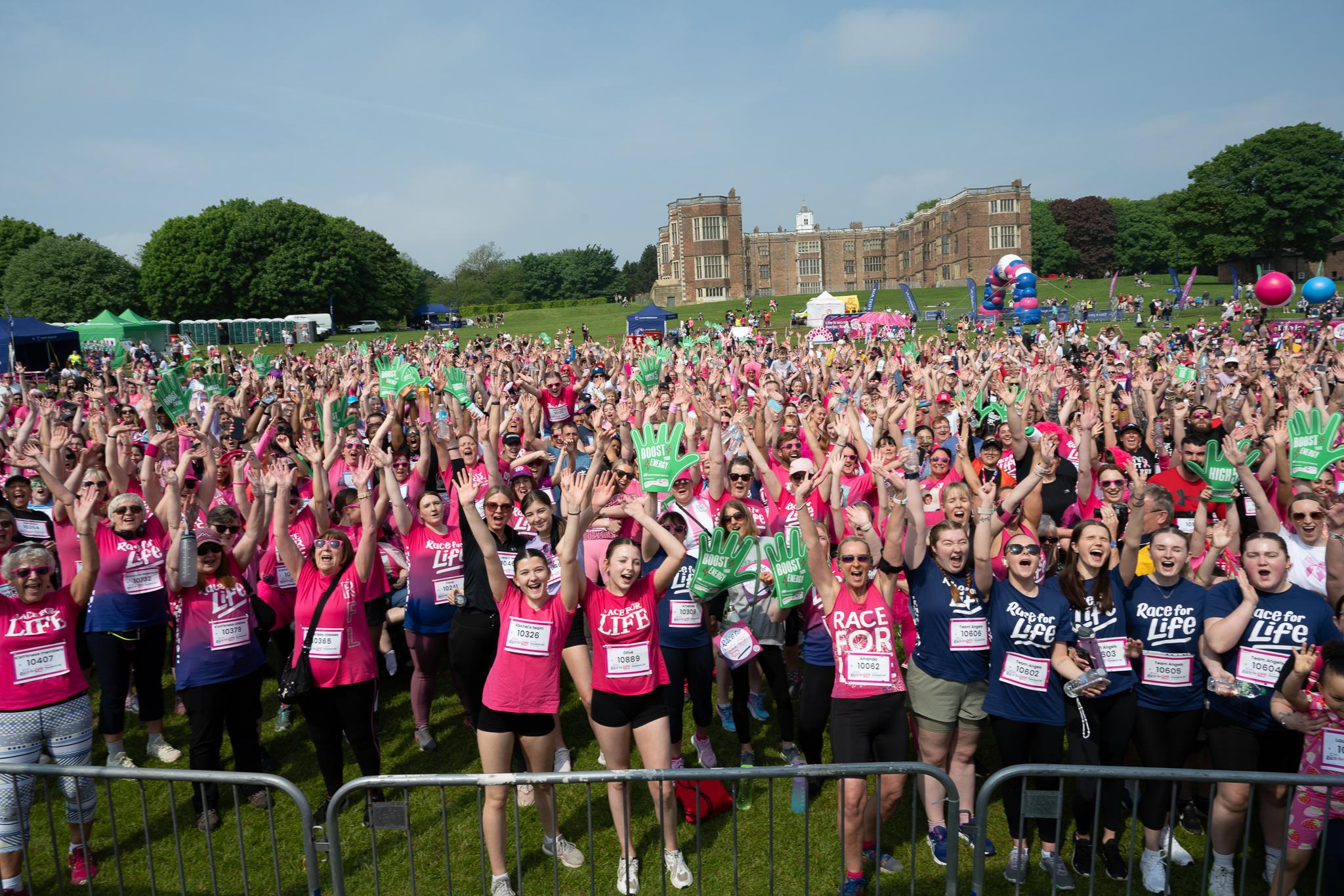 A large crowd of runners at a Race for Life event are standing outside with their hands in the air in celebration.