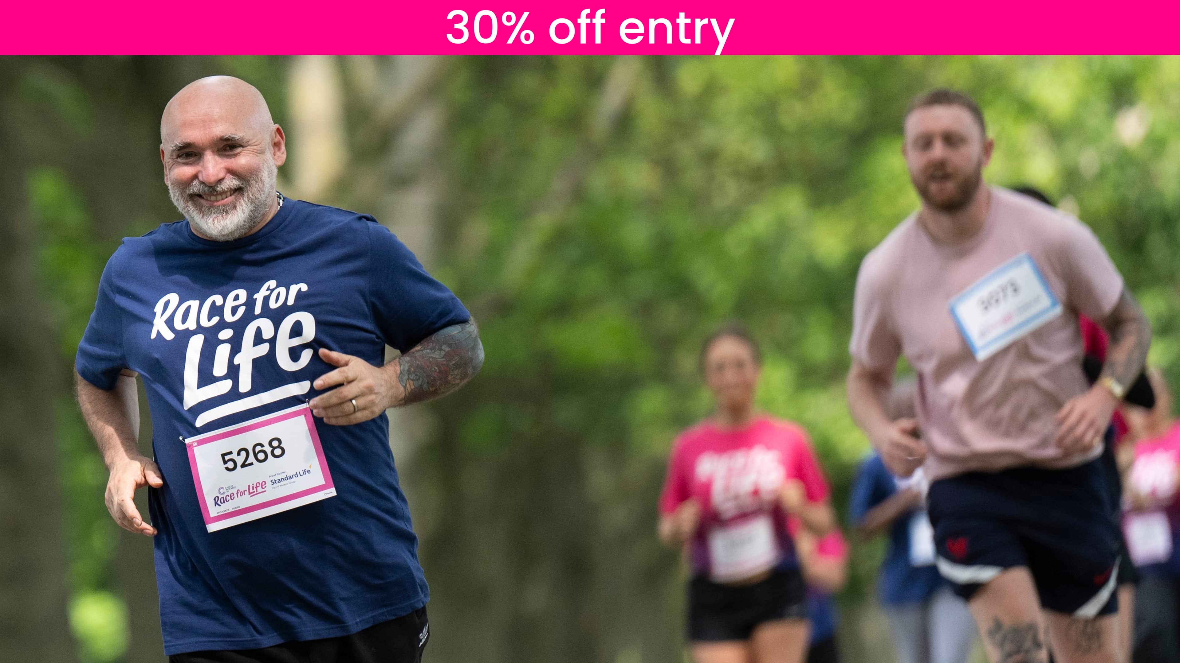 A man is running in park and wearing a blue T-shirt, while taking part in a Race for Life 10K event. He looks happy.