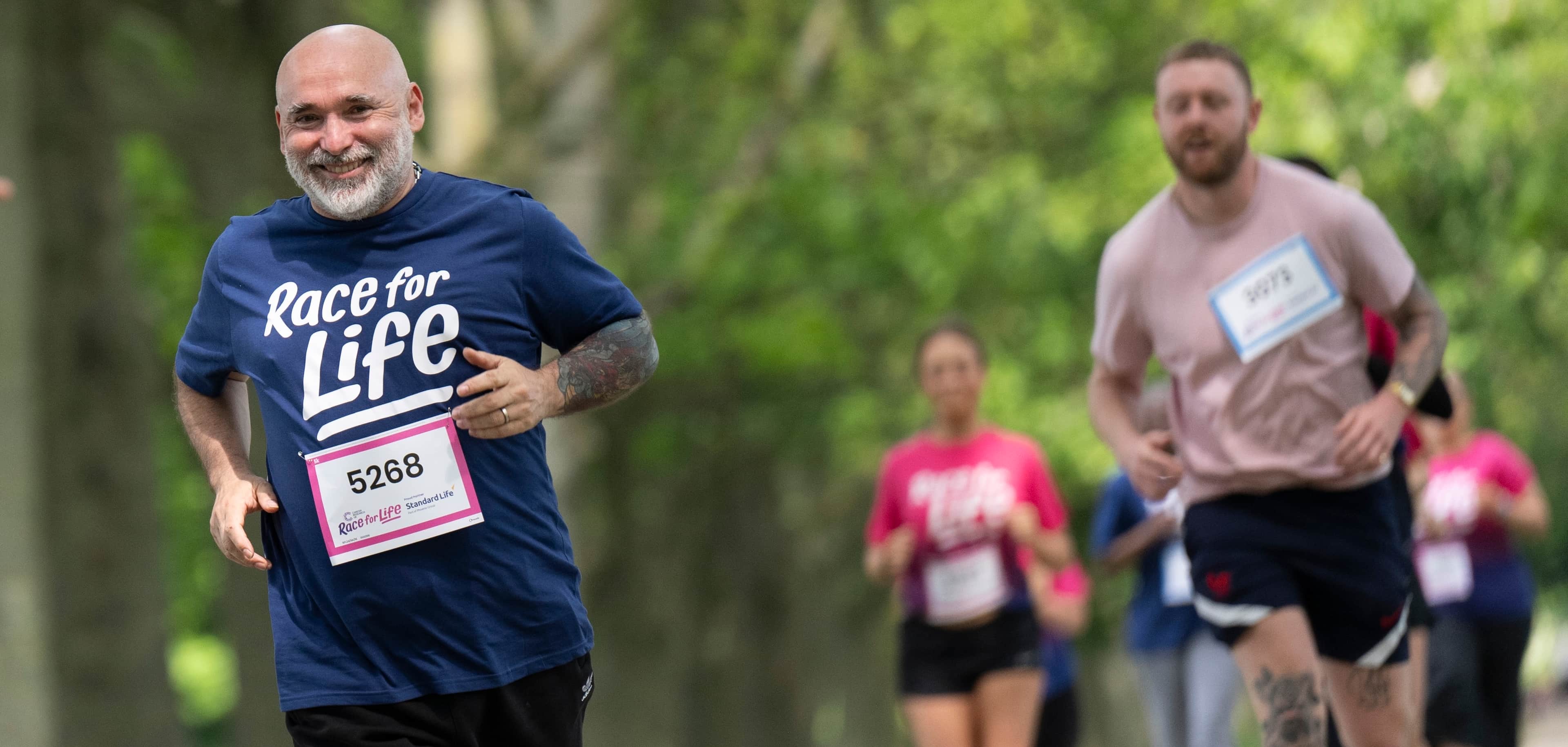 A man is running in park and wearing a blue  T-shirt, while taking part in a Race for Life 10K event. He looks happy.