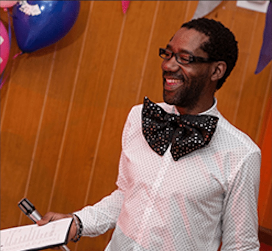 A smiling man wearing an oversized bowtie is standing in front of balloons and a chalkboard sign that says quiz night.