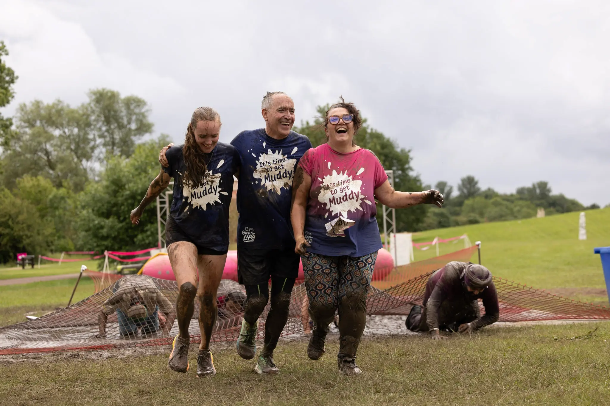 Three people wearing Race for Life t-shirts walk in front of a muddy obstacle course.