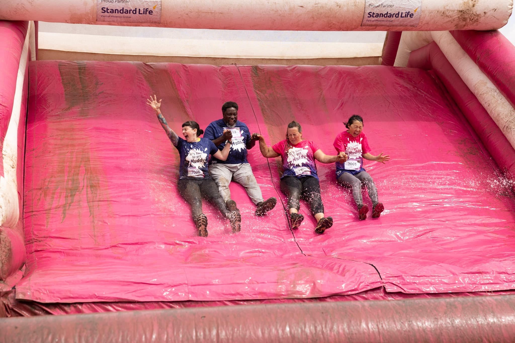 Four adults going down a Pretty Muddy slide, smiling and laughing.