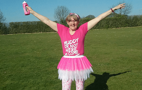 A woman standing in a field wearing a pink tutu and holding a pink water bottle. Her t-shirt reads Muddy or not here we come.