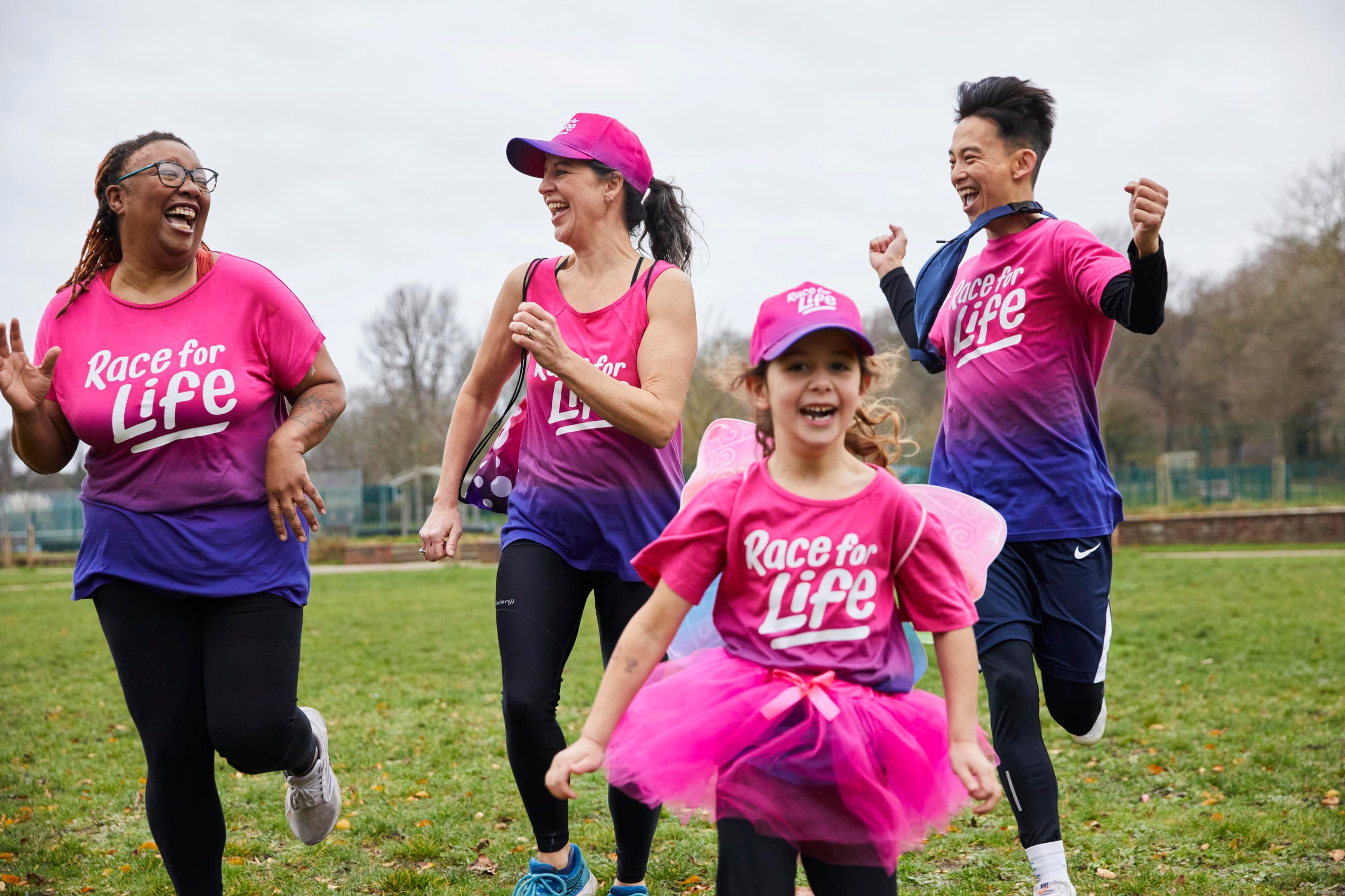 Two women, a man and a young girl in Race for Life branded t-shirts are laughing and running in a park.