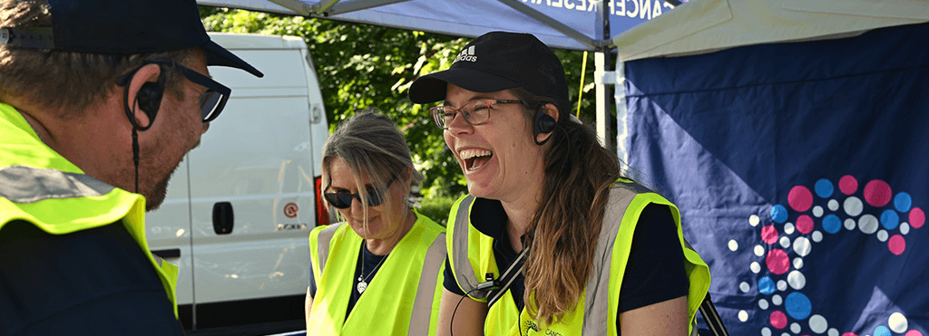 Event volunteers wearing Hi-Vis vests and earpieces, laughing as they prepare the enquiries tent for the Race for Life event.