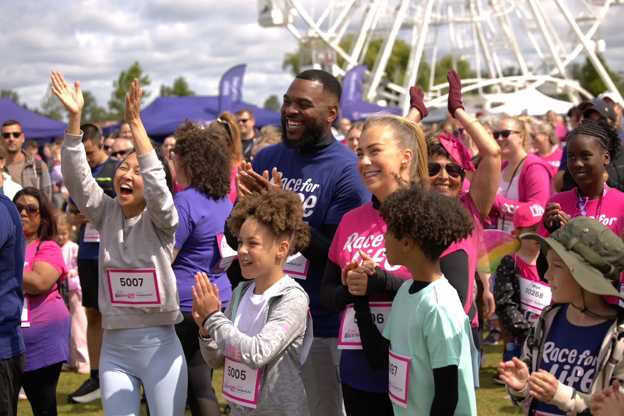 Two adults and two children wearing Race for Life t-shirts, standing in a crowd and clapping.