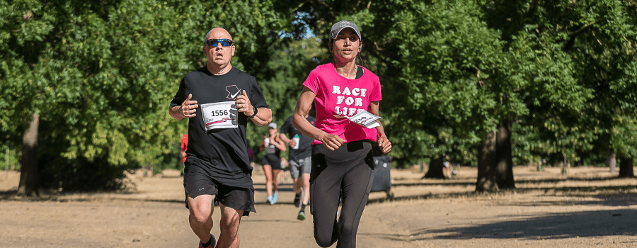 A man in a black t-shirt and woman in a pink Race for Life t-shirt running through a park