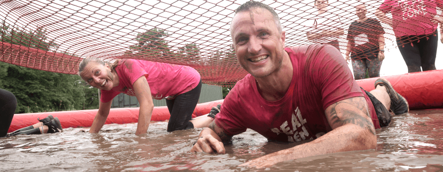 A man and a woman wearing pink t-shirts crawling through a big puddle of mud