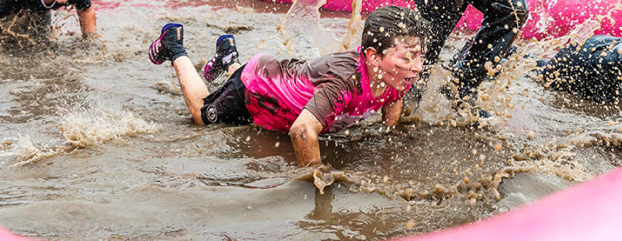 A boy in a pink t-shirt crawling through a muddy puddle